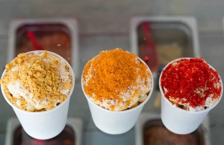 Esquites en vasos con distintas coberturas frente a una barra